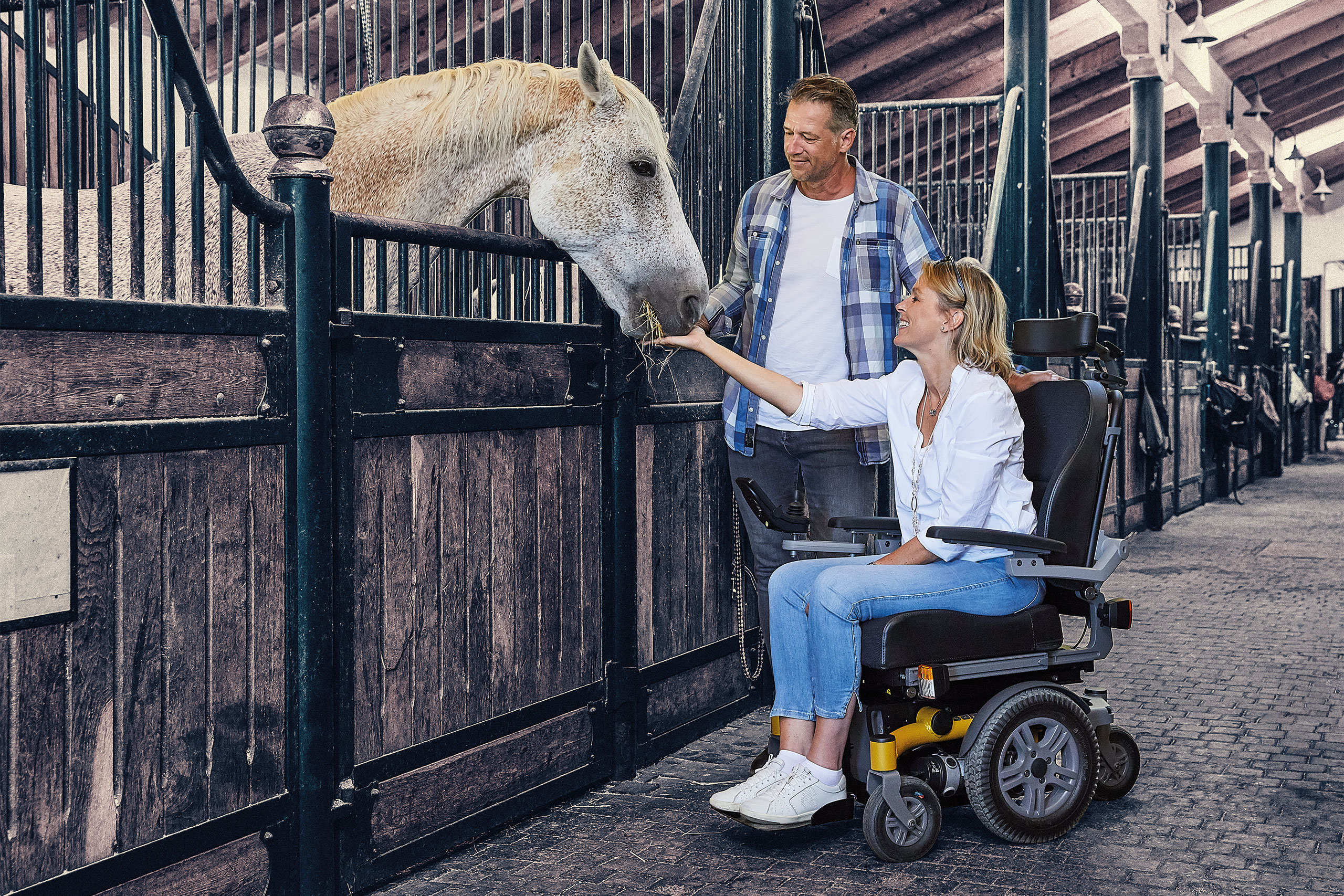 Middle-aged lady, accompanied by her partner, stands in front of a horse box in a large stable with her SANGO slimline powered wheelchair and gives the horse carrots to nibble on.