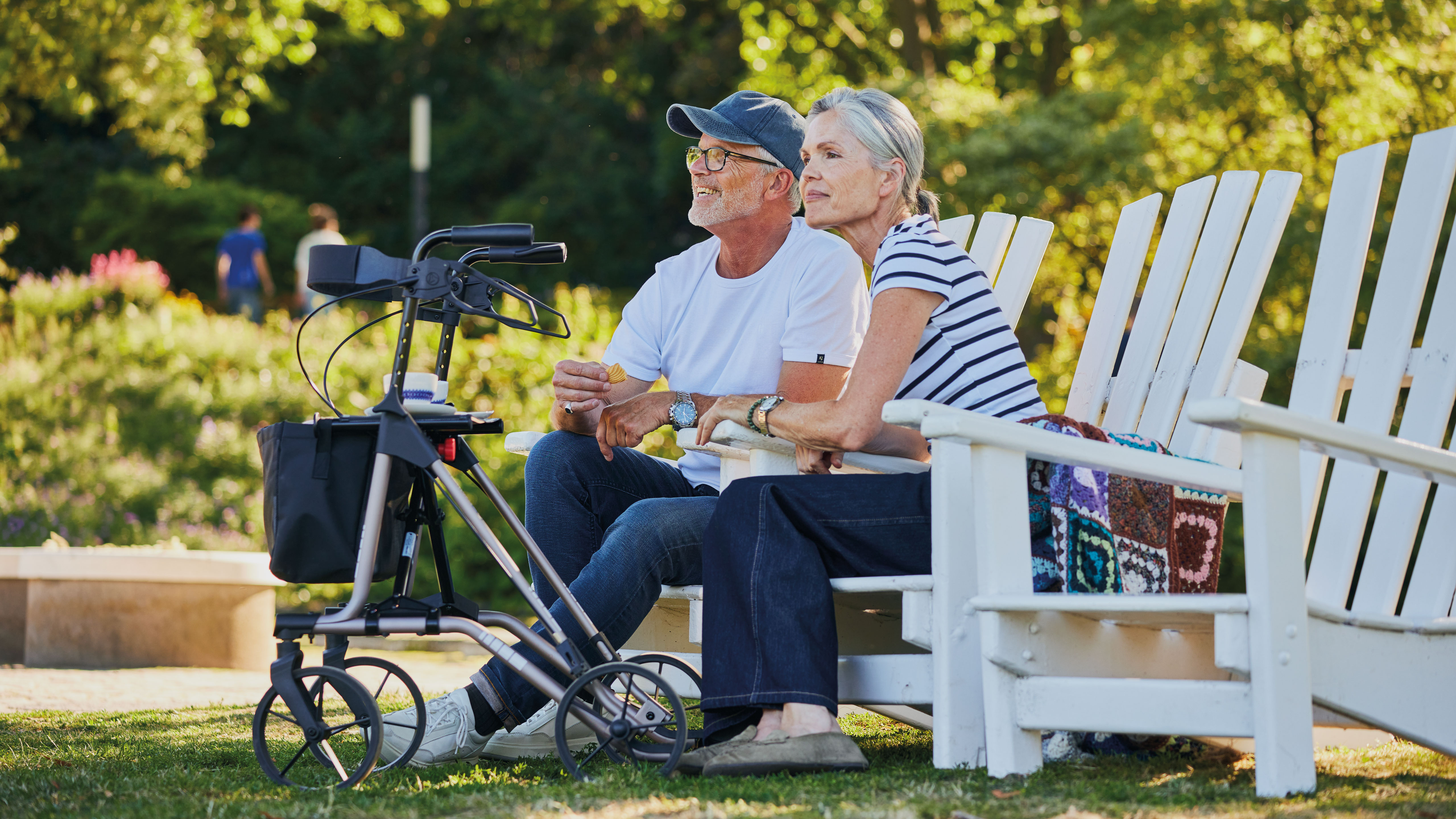 TAiMA M-E Eine ältere Frau und ein älterer Mann sitzen an einem sonnigen Tag auf einer Bank im Park und blicken gemeinsam ins Grüne. Vor ihnen steht ein Rollator TAiMA M-E in Terrarosa.
