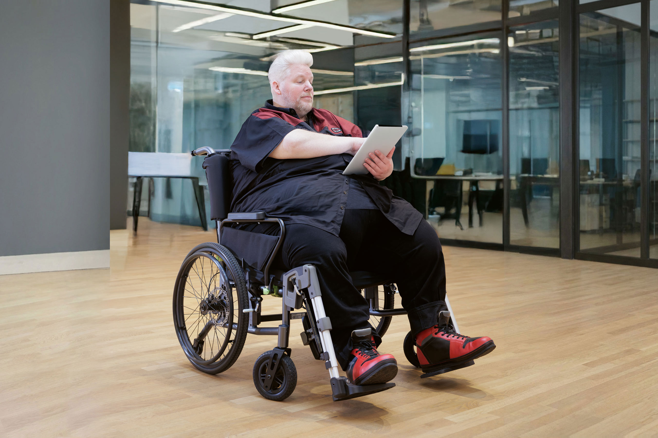 Sehr korpulenter Mann mittleren Alters in seinem manuellen XXL-Rollstuhl tauron|rsi steht in einer modernen Bürofläche und liest in seinem Tablet.
